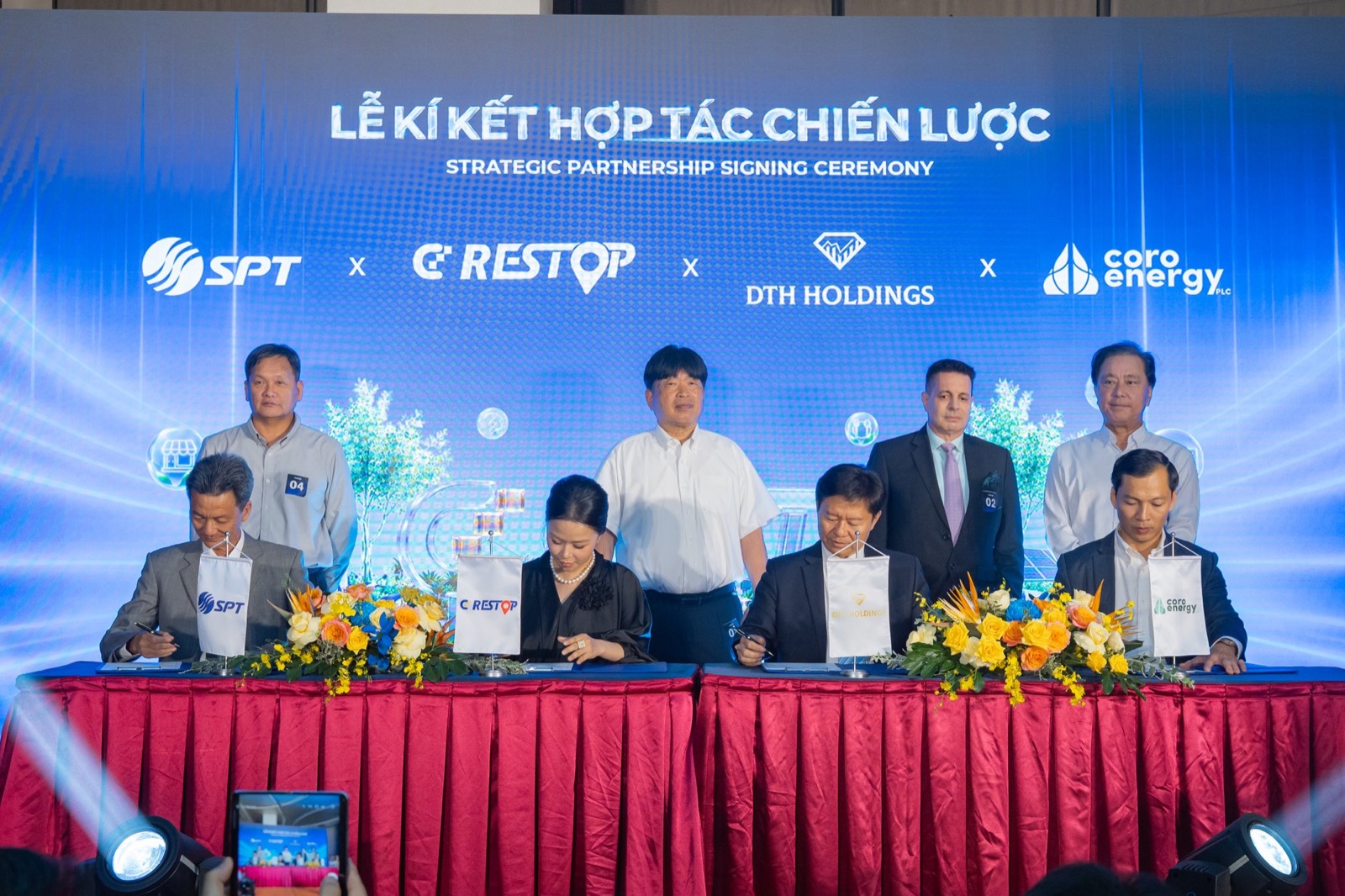 Bốn đại diện ngồi ký văn kiện tại bàn phủ khăn đỏ, trước mặt là cờ để bàn của Tập đoàn SPT, CD Restop, DTH Holdings và Coro Energy, phía sau là các đại biểu chứng kiến