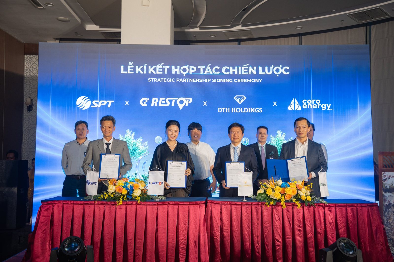Представители Группы SPT, CD Restop, DTH Holdings и Coro Energy держат только что подписанный документ, стоя за столом для подписания под красной скатертью, на LED-экране — логотипы четырёх сторон