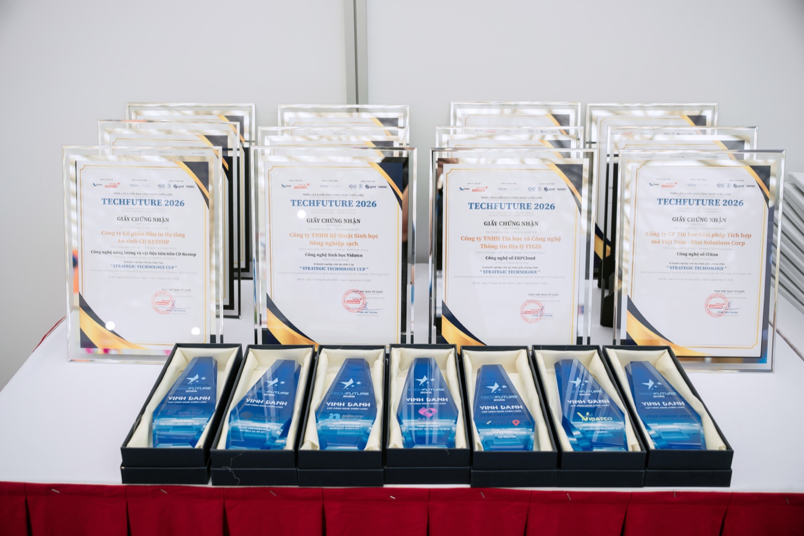 Bàn trưng bày các giấy chứng nhận TechFuture 2026 trong khung mica cùng dãy cúp pha lê xanh khắc chữ Vinh danh – Cúp Công nghệ Chiến lược, giấy ngoài cùng bên trái ghi tên Công ty Cổ phần Đầu tư Hạ tầng An sinh CD Restop